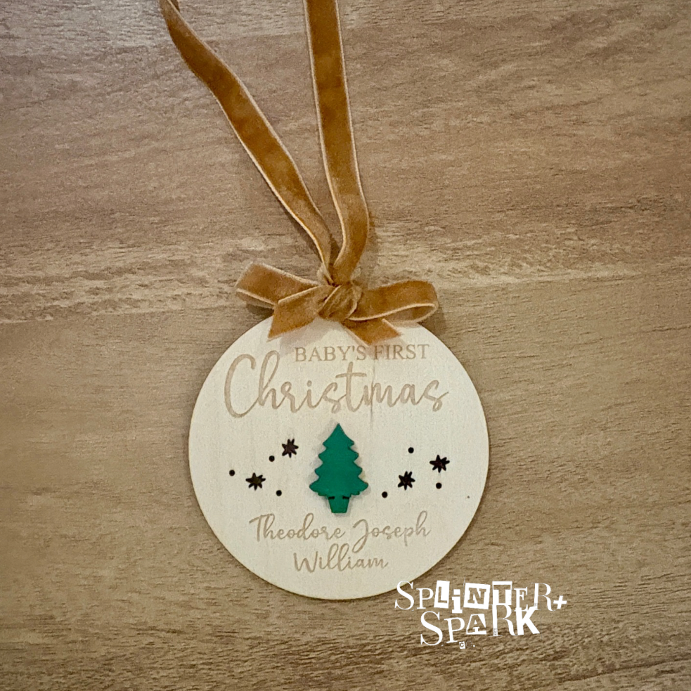 Round wooden ornament with 'Baby's First Christmas' text and tree design on a wooden surface, branded 'Splinter + Spark'.