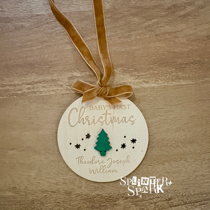 Round wooden ornament with 'Baby's First Christmas' text and tree design on a wooden surface, branded 'Splinter + Spark'.